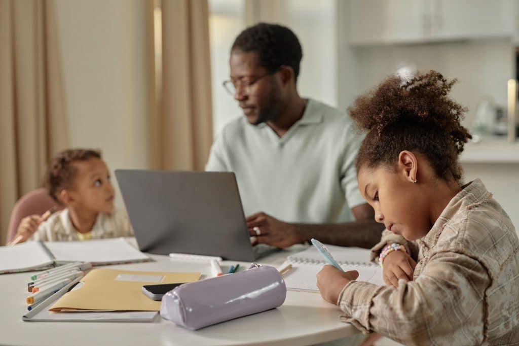Smart Little Black Girl Writing in Notebook while Studying at Table with Family at Home